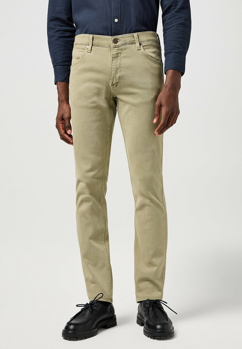 Homme portant un pantalon beige, des chaussures noires à lacets et une chemise bleu foncé à manches longues, debout devant un fond uni.