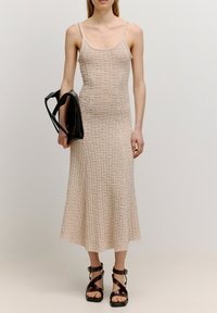 Strick-Midikleid in Hellbeige mit strukturiertem Karomuster. Dünne Träger, taillierter Oberteil, ausgestellter Saum. Modell hält eine schwarze Handtasche. Braune Sandalen mit Riemchen.