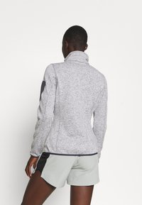 Persoon met kort haar in een grijze gestructureerde jas met elleboogstukken en lichtgrijze shorts, staande tegen een witte achtergrond.