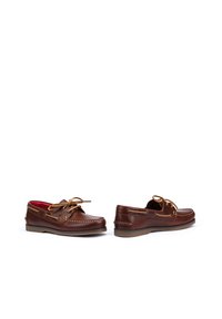 Los zapatos de barco de cuero marrón presentan un diseño cosido, suela de goma y forro de tela con un acento rojo. Incluyen un detalle de cordones en los lados.