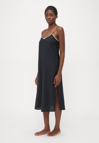 Kvinne iført en svart midikjole i silk, med hvite prikker, tynne stropper og splitt i siden, som står barbent mot en ensfarget bakgrunn.