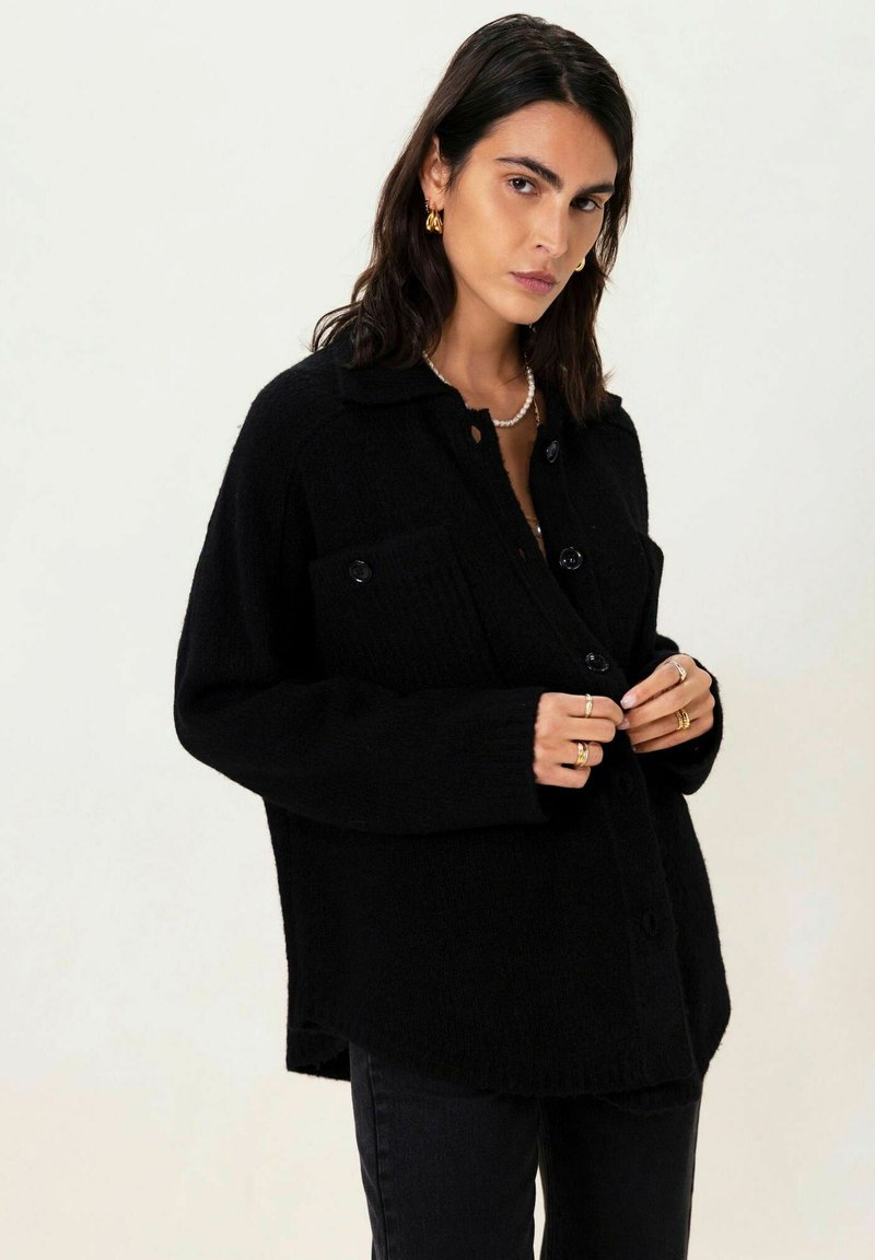 Cardigan noir oversize à boutons en tissu texturé, avec un col, des poches frontales et une coupe loose. Porté avec un jean sombre et des bagues.