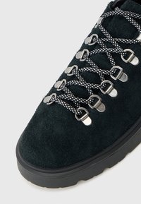 Sapatilha de caminhada em camurça negra com bico arredondado, apresentando cordões com padrão preto e branco e ilhós metálicos, assente sobre uma sola de borracha texturizada preta.