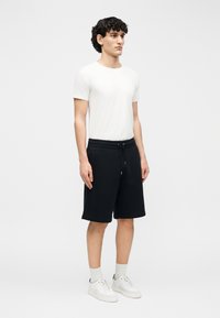 Joven de pie, vestido con una camiseta blanca, pantalones cortos negros con cordón, calcetines blancos y zapatillas blancas, sobre un fondo claro liso.