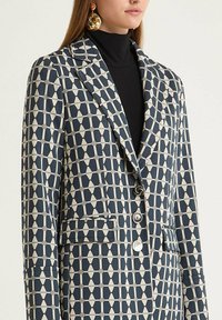 Blazer à motif géométrique bleu marine et crème, avec un col à revers cranté, une fermeture à un bouton et des poches latérales. Texture de tissu lisse.