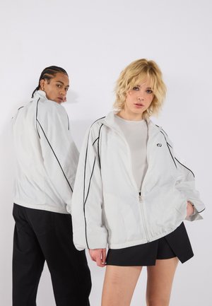 White zip-up jacket with black accents, featuring a high collar and relaxed fit. Includes elastic cuffs and hem for adjustable sizing.