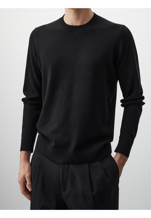Mand iført en ensfarvet sort langærmet rundhals sweater med ribkanter ved ærmer og kant, kombineret med sorte bukser, hænder afslappede.