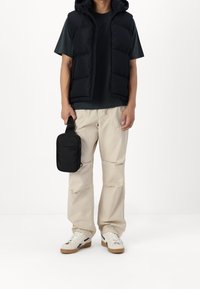 Czarna kamizelka z ociepleniem i kapturem, ciemnozielona koszulka, beżowe spodnie, białe trampki z brązowymi elementami oraz mała czarna torba na ramię.