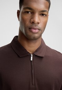 Braunes Zip-Polo-Shirt mit Kragen, das sich durch eine glatte Stofftextur und einen feinen Glanz auszeichnet. Betont durch ein kleines silbernes Reißverschlussdetail.