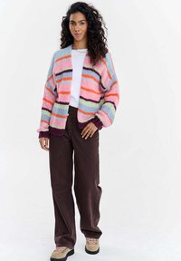 Žena v mnohobarevném pruhovaném pleteném kardiganu, bílém tričku, tmavě hnědých širokých kalhotách a béžových turistických botách, stojící na bílém pozadí.