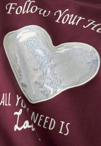 Bordowy sweter z dużą, srebrną, błyszczącą aplikacją w kształcie serca. Napis brzmi: „Podążaj za swoim sercem” oraz „Wszystko, czego potrzebujesz, to miłość” na białym tle.