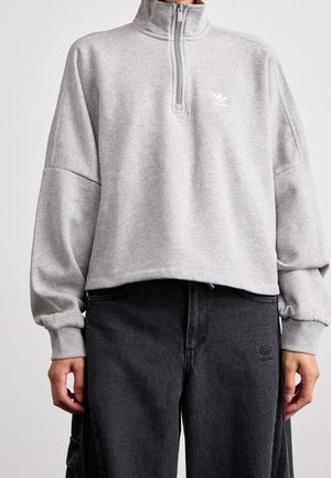 Hellgraues Sweatshirt mit Viertel-Reißverschluss und hohem Kragen, getragen mit schwarzen Adidas-Hosen, die Logos auf der Brust und am Oberschenkel zeigen.