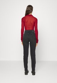 Mujer con cabello largo castaño, vistiendo una blusa roja transparente de manga larga, unos pantalones vaqueros negros de tiro alto y botas negras, de pie frente a una pared blanca.