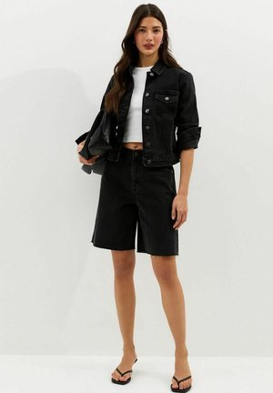 Mujer joven con chaqueta de mezclilla negra, pantalones cortos negros hasta la rodilla, top corto blanco, sandalias negras, sosteniendo un bolso de mano negro contra un fondo blanco.