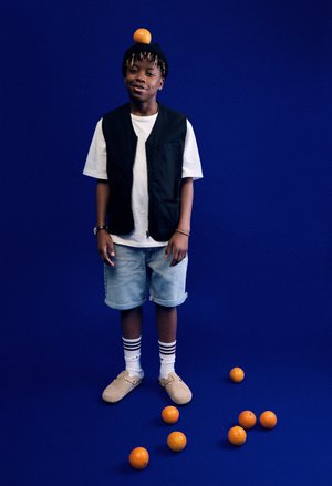 Black vest over white shirt, denim shorts, striped socks, brown slip-on shoes. Orange on head, surrounded by oranges on blue background.