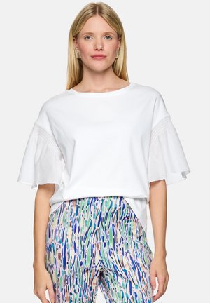 Blonde Frau trägt weißes, kurzärmeliges, gerafftes Oberteil und gemusterte Hosen in Blau, Grün und Beige, stehend vor einem schlichten Hintergrund.