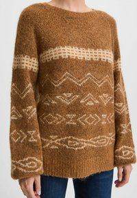 Maglione marrone lavorato a maglia con dettagli crema a motivo, caratterizzato da disegni geometrici. Le maniche lunghe e la vestibilità rilassata mettono in risalto la sua superficie testurizzata.