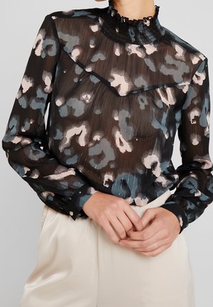 Blouse noire transparente à manches longues avec un motif floral abstrait beige et gris, col montant à volants, et pantalon beige. Mains jointes devant.