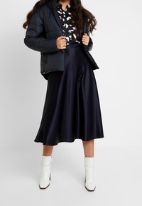 Doudoune marine avec capuche, blouse à motifs noir et blanc, jupe fluide en satin noir et bottines blanches à fermeture éclair sur la cheville avec un talon carré.