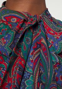 Färgglad blus med paisleymönster i rött, blått och grönt. Har knytband i halsen och strukturerat tyg. Närbild som framhäver designdetaljerna.