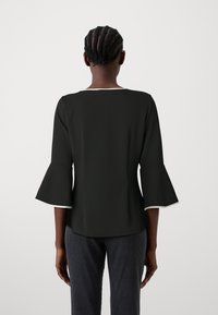 Černý top s rukávy do zvonu, kulatým výstřihem a bílým lemem na okrajích rukávů, v kombinaci s žebrovanými šedými kalhotami.