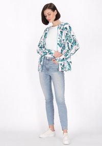 Blomstret zip-up jakke med grønne blade og lyserøde blomster, parret med tætsiddende lyseblå jeans og hvide sneakers, præsenterende et afslappet look.