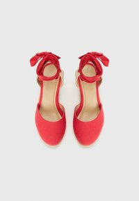 Röda espadrilloskor med en slät tygövre, öppen häl och dekorativa röda snören runt vristerna, med en vävd jutesula.