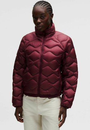 Donkere persoon draagt een mahoniegekleurde gewatteerde pufferjas met rits aan de voorkant en hoge kraag, gecombineerd met lichtgekleurde broek.