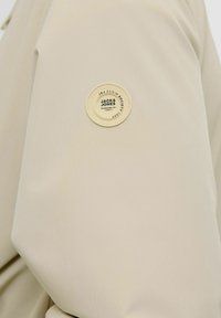 Veste à capuche en tissu beige avec un patch circulaire portant l'inscription "JACK & JONES" sur la manche gauche. Texture lisse et design simple.