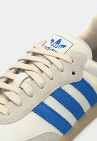 Adidas Samba sneakers i kremfarget skinn med blå semskede striper, beige snørebånd og en teksturert såle. Har logo og modellnavn på tungen.