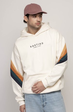 White hooded sweatshirt with black logo, featuring orange and teal stripes on sleeves, paired with light blue jeans and a pink cap.