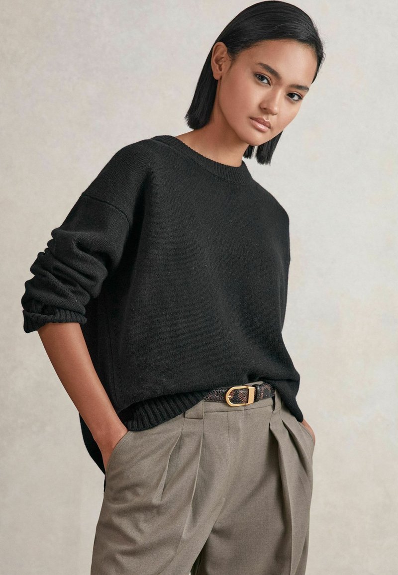 Pull en tricot noir à manches retroussées, associé à un pantalon gris plissé et une ceinture texturée avec une boucle en or. Design simple et moderne.