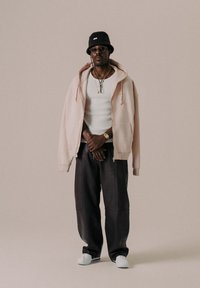 Haut de hoodie zippé rose clair, débardeur blanc, pantalon noir ample et chaussures blanches. Porte un chapeau en seau noir et des lunettes, avec des colliers superposés.