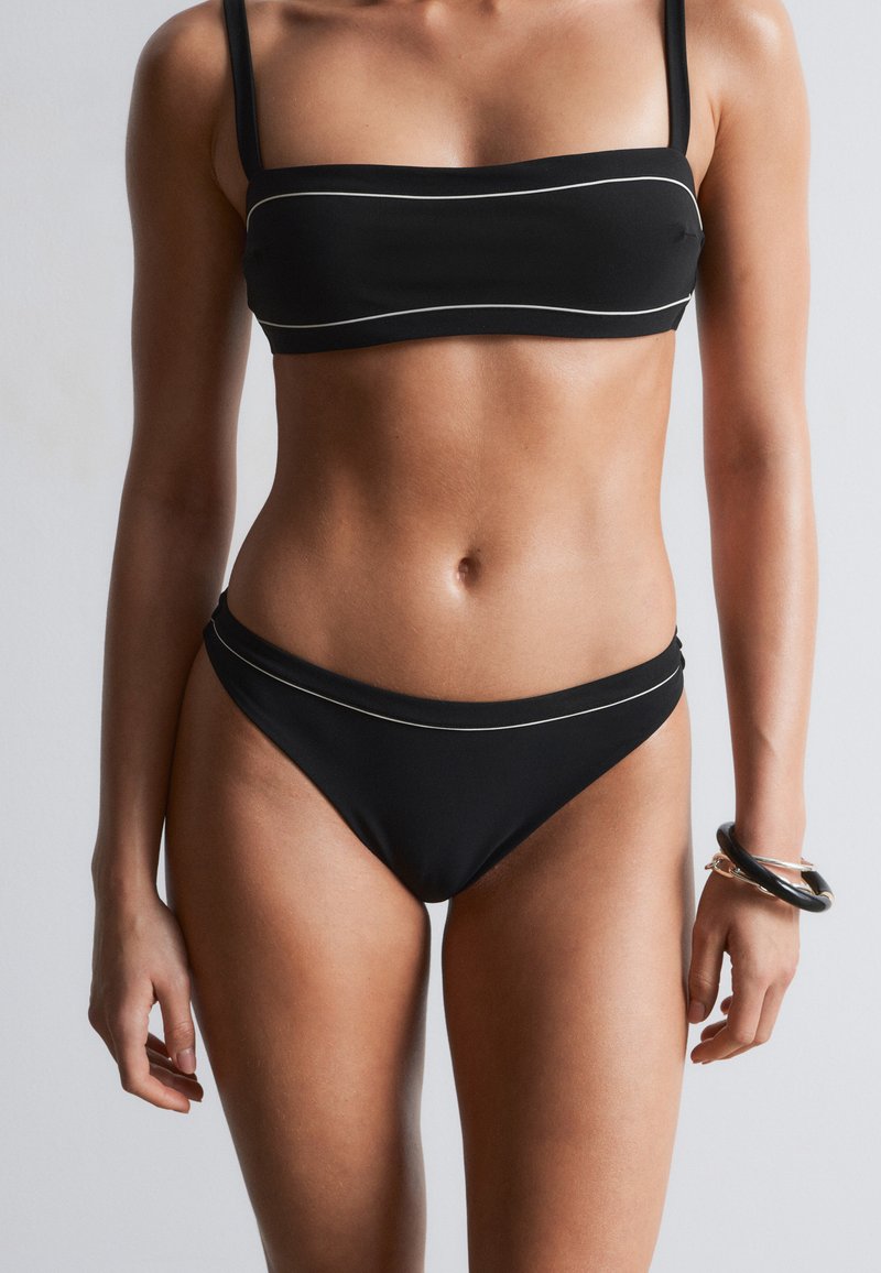 Femme portant un maillot de bain deux pièces noir avec des finitions blanches fines, debout devant un fond gris neutre, portant des bracelets noirs au poignet droit.
