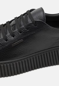 Baskets en cuir noir avec dessus texturé, lacets plats noirs et semelle extérieure en caoutchouc noir épaisse avec des rainures verticales.