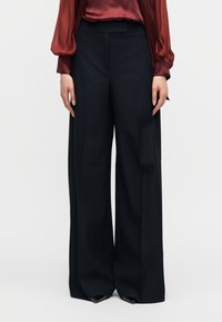 Μαύρο παντελόνι με φαρδιά πόδια συνδυασμένο με διαφανές μπορντό μπλούζα με μακριά μανίκια, χέρια χαλαρά στο πλάι, στέκεται σε απλό λευκό φόντο.