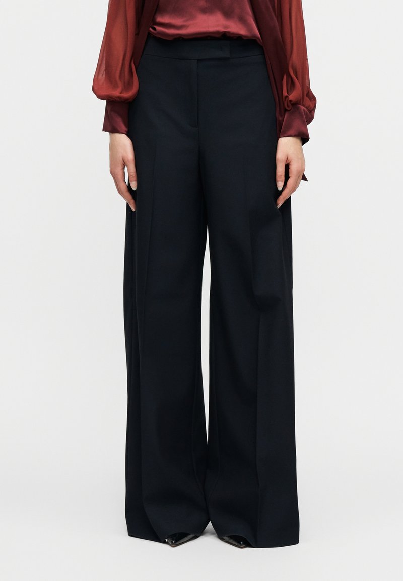 Μαύρο παντελόνι με φαρδιά πόδια συνδυασμένο με διαφανές μπορντό μπλούζα με μακριά μανίκια, χέρια χαλαρά στο πλάι, στέκεται σε απλό λευκό φόντο.