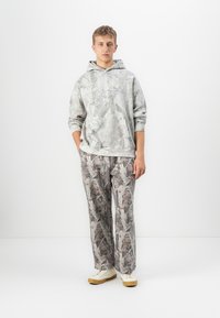 Giovane uomo in piedi su uno sfondo bianco, indossa un hoodie grigio a fantasia, pantaloni larghi coordinati e sneakers bianche con suola in gomma.