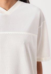 Haut blanc à manches courtes présentant une section supérieure texturée à côtes et un panneau en maille en dessous avec des perforations régulièrement espacées.
