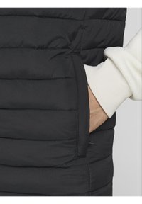 Gilet imbottito nero con cuciture orizzontali, tasche laterali e polsini a coste crema visibili. Texture liscia, design funzionale.