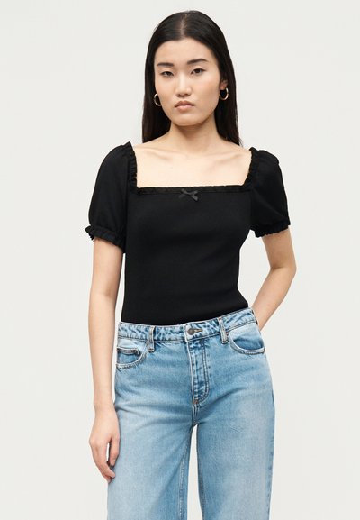 Νεαρή γυναίκα με μακριά σκουρόχρωμα μαλλιά, φορώντας μαύρο τοπ με φουσκωτά μανίκια και τετράγωνη λαιμόκοψη, καθώς και ανοιχτό μπλε ψηλόμεσα τζιν, στέκεται μπροστά σε απλό φόντο.