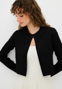 Maglione nero con scollatura rotonda, caratterizzato da decorazioni di perline sulle spalle. Il tessuto è morbido e ha un design corto e aderente.