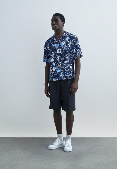 Homme portant une chemise à manches courtes bleu marine à motifs floraux, un short noir, des chaussettes blanches et des baskets blanches, debout devant un fond uni.