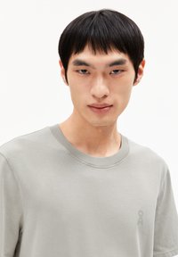 Jonge man met kort zwart haar, gekleed in een lichtgrijze t-shirt met ronde hals en een klein logo op de borst, tegen een witte achtergrond.