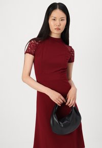 Rochie burgundă cu mâneci scurte din dantelă și corset structurat. Modelul ține o geantă neagră, cu textura moale și mânere rotunjite.