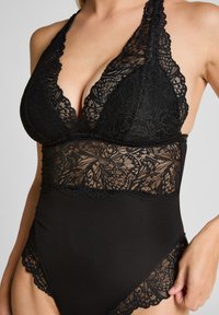 Svart spetsbody med djup urringning, scallopade kanter och blommig spetsdetalj vid midjan och benöppningarna. Strukturerad tyg.