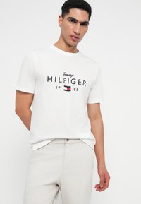Mladý muž oblečený v bílém tričku Tommy Hilfiger 1985 a béžových kalhotách, stojící na světlém jednoduchém pozadí a dívající se do kamery.