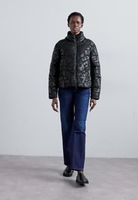 Zwarte pufferjack met een geometrisch patroon, hoge kraag en ritssluiting; gecombineerd met blauwe flare jeans en zwarte schoenen.