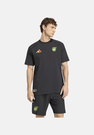 Hombre vistiendo camiseta deportiva negra de manga corta y pantalones cortos con los logotipos verdes y amarillos del equipo de fútbol de Jamaica sobre un fondo liso.