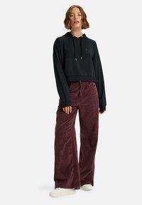 Czarny skrócony hoodie z kieszenią z przodu i kapturem na sznurki, zestawiony z szerokimi bordowymi spodniami z korduroju i białymi trampkami.